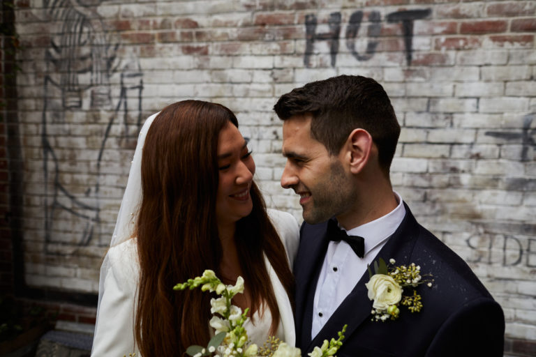 Wedding Photographer, couple looking at each other and smiling with graffitied brick wall behind them
